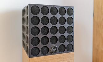 A modern speaker with a square shape and a black, perforated surface. The speaker stands on a wooden base and has a simple design.