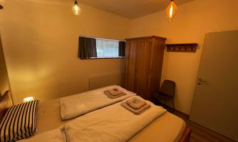 A cozy bedroom with two beds, a wooden wardrobe, and warm light sources. The walls are bright, and the room feels inviting.