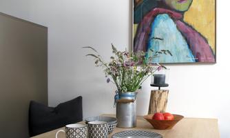 A stylish dining table with various cups, a bowl of apples, and a floral arrangement. In the background, a colorful painting hangs.