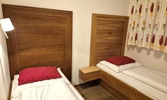 Two single beds in a room with wooden beds and red pillows. A window with colorful curtains brings some light into the room.