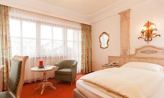 A cozy hotel room with a large window and warm colors. There is a bed, a table with a chair, and a decorative wall.