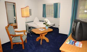 A cozy hotel room with a bed, a table, and an armchair. The room has bright curtains and a plant on the table.