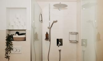 A modern shower with glass partitions and a large rain showerhead. In the background, items like soap and toilet paper are neatly arranged.