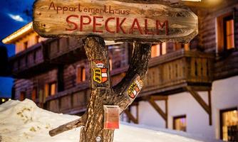 A wooden sign with the inscription "Appartementhaus SPECKALM" stands in the snow. In the background, alpine buildings and a blue sky are visible.