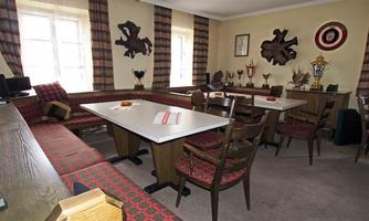 A cozy room with tables and chairs, adorned with trophies and pilgrimage items. The windows are decorated with checkered curtains, and the floor is covered with carpet.