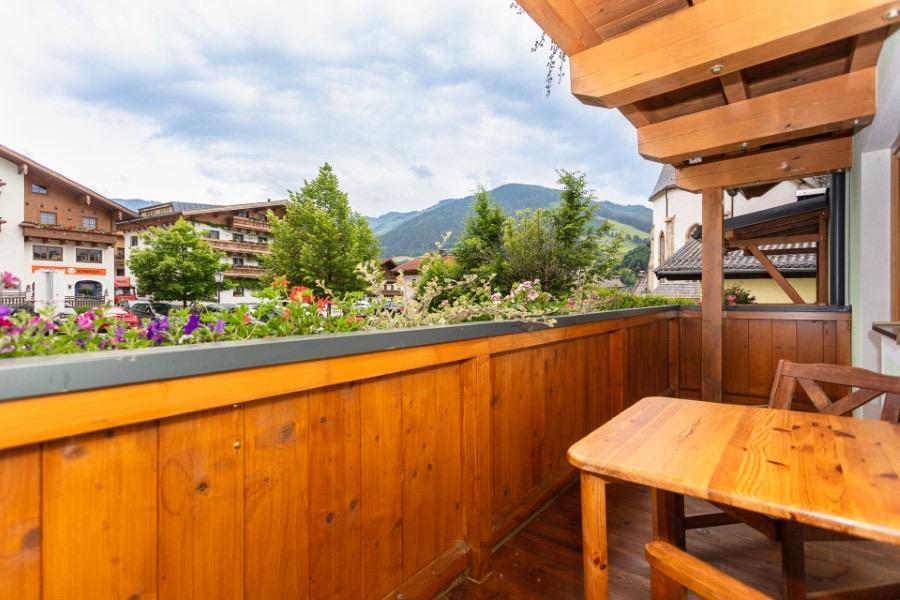 A balcony with a wooden railing and a table. In the background, trees and mountains can be seen, the sky is cloudy.