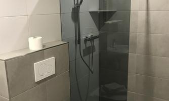 A modern bathroom with a glass shower wall and gray tiles. Pink towels are placed on the toilet.