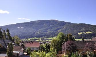 A picturesque landscape with a green mountain in the background. In the foreground, typical houses and trees can be seen.