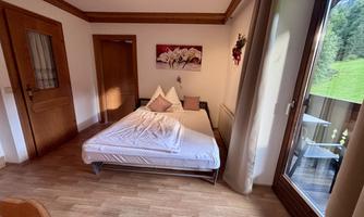 A simple room with a bed and wooden details.  
Large windows offer a view of the greenery.
