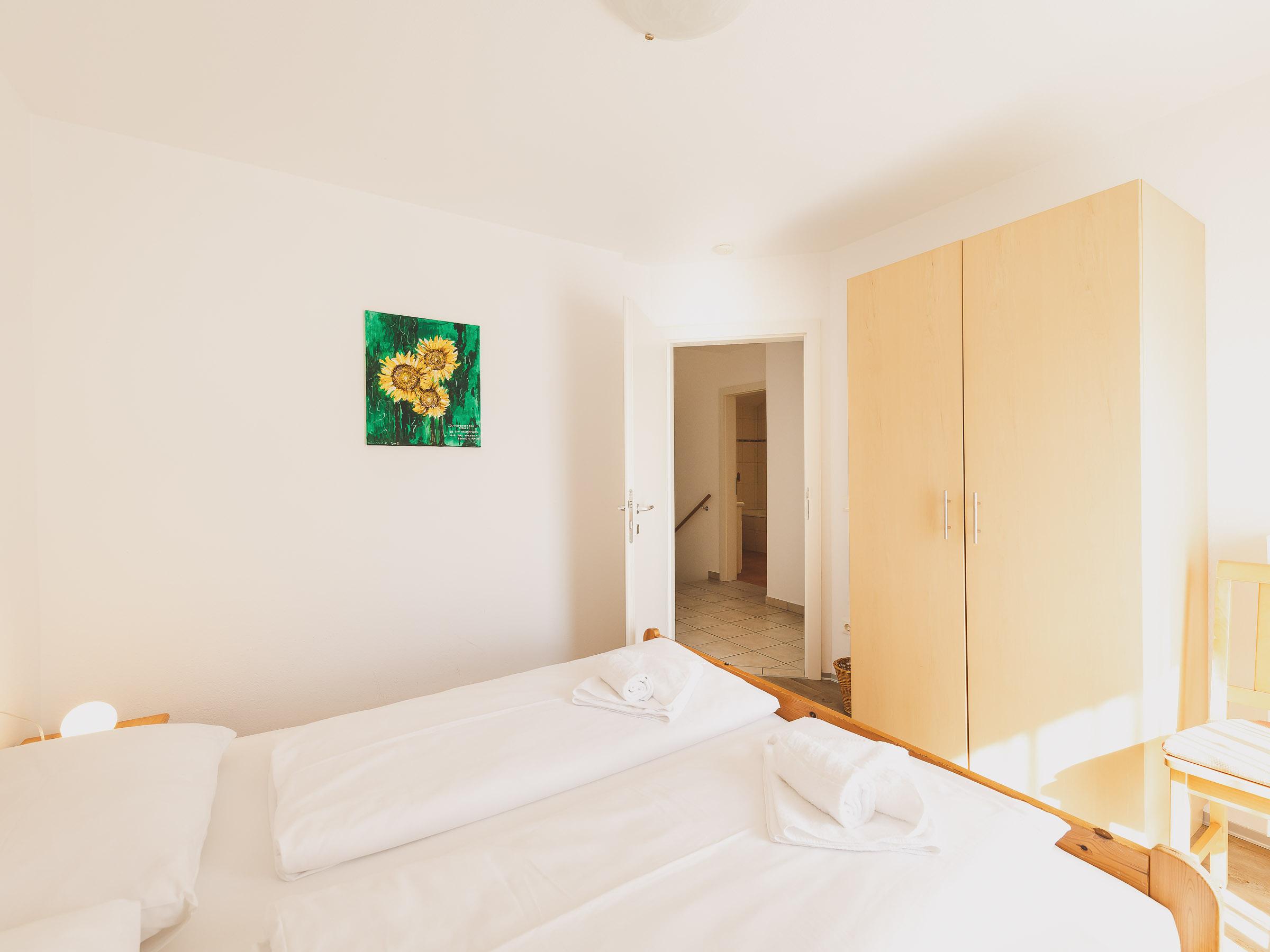 A bright and modern bedroom with two beds and a large wardrobe. A painting of sunflowers hangs on the wall.