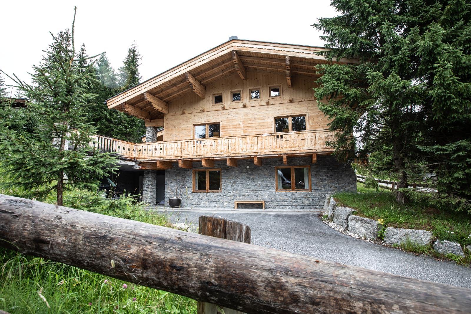 A beautiful wooden house with a large balcony amidst spruce trees. The facade combines wood and stone and is idyllically situated in nature.