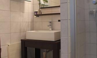 A modern bathroom with a white sink and a shower. The walls are tiled, and a small bouquet of flowers is on the shelf.