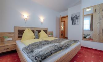 A cozy bedroom with a large bed and yellow pillows. The walls are light, and there is a mirror as well as a wooden dresser.