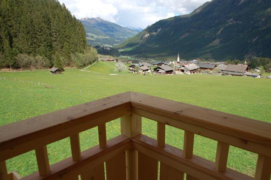A beautiful view from a balcony overlooking a green meadow and a small village in the mountains. In the background, high mountains and a blue sky can be seen.