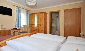A bright bedroom with a double bed, wooden furniture, and a small table. The room has a window with curtains and an adjoining bathroom.