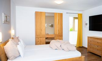 A bright bedroom with a double bed and a wooden dresser. The walls are in neutral colors, and a large TV is mounted opposite the bed.