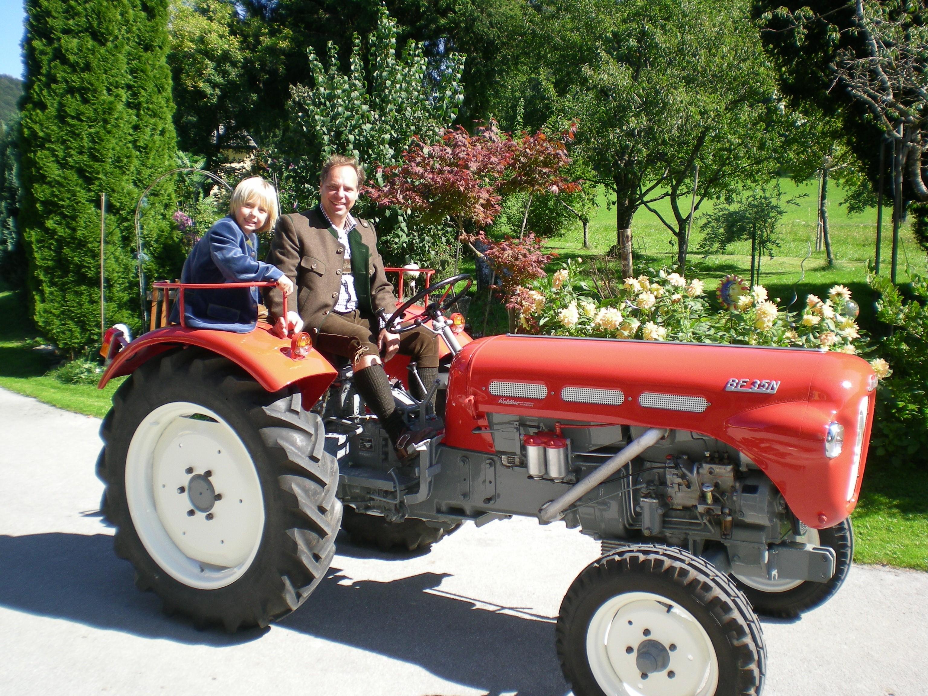 A red tractor is standing on a path where two people are sitting. In the background, there are green trees and colorful flowers.