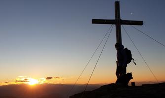 A hiker stands silhouetted before a cross on the summit. The sun rises on the horizon, bathing the scene in golden light.