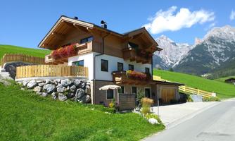 A charming mountain house with a wooden veranda and colorful flowers. In the background, the majestic mountains and a clear sky are visible.
