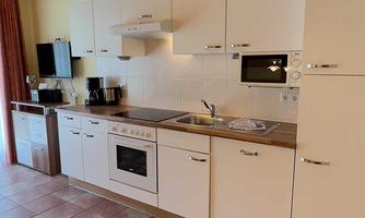 A modern kitchen with white appliances and wooden shelves. The room is bright and inviting, with tiled flooring and a pleasant atmosphere.