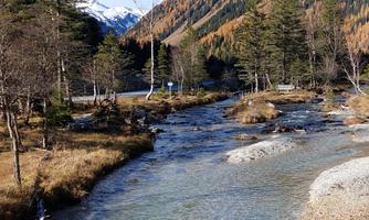 A clear river flows through a picturesque landscape with tall mountains and trees. The sky is blue and the mountains are partially covered with snow.