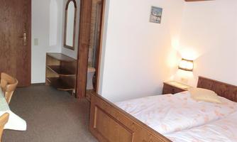 A cozy room with two beds, a table, and wooden shelves. The walls are painted light, and there is a bedside lamp.