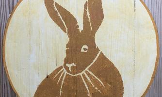 A brown rabbit is depicted in a round design on a wooden background. The rabbit has large ears and a round body.