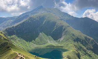 A stunning mountain landscape with a clear blue lake and green meadows. The sky is partly cloudy and the mountains are majestic.