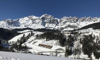 A majestic mountain landscape with snow-capped peaks and a clear blue sky. In the foreground, there are snow-covered hills and some wooden chalets.