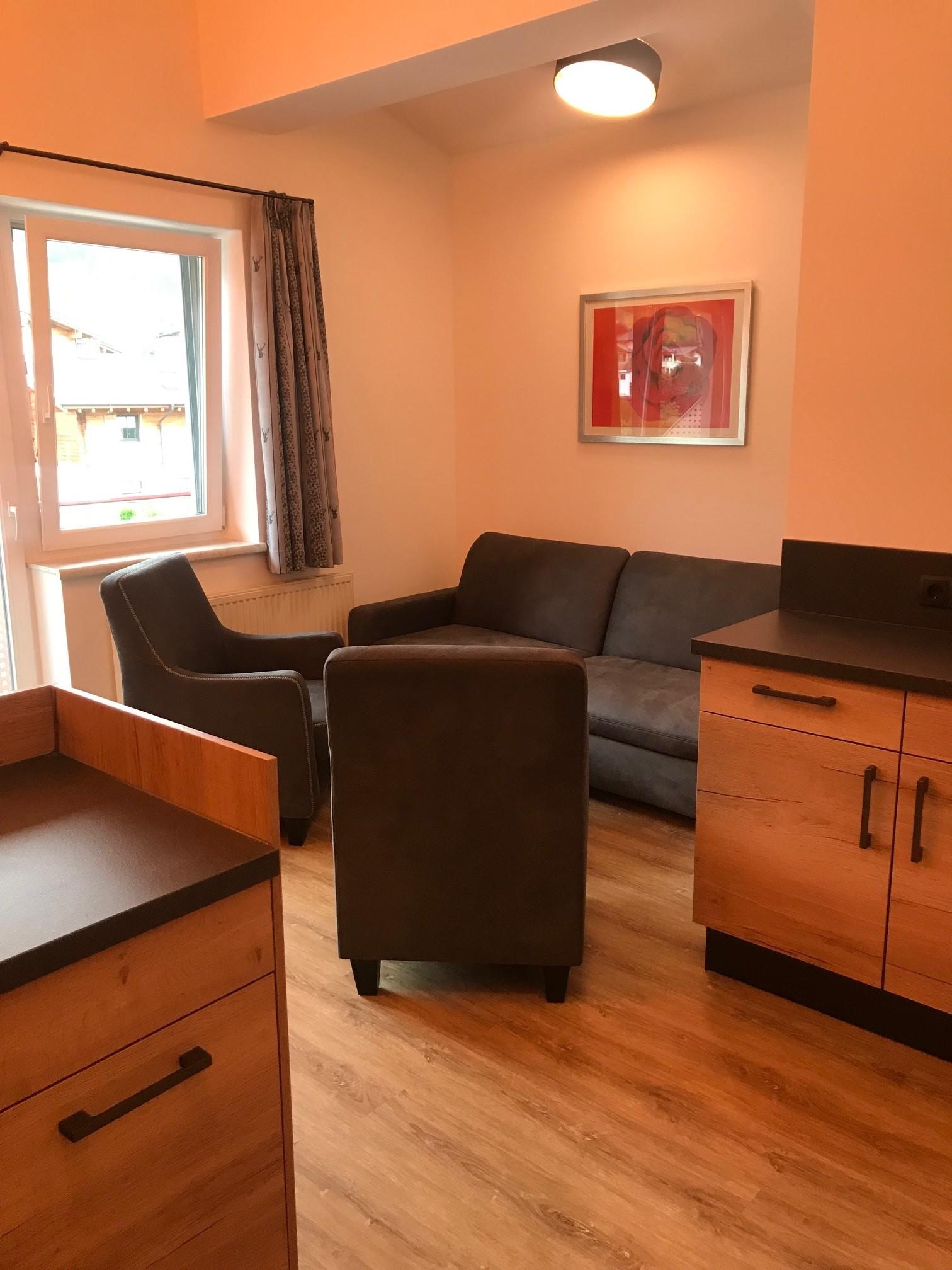 A cozy living room with a gray sofa and an armchair. There is a small table and a bright window front.
