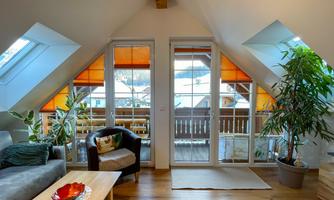 A bright attic apartment with large windows and orange blinds. The room features plants and cozy furniture.