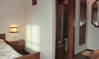 A simple bedroom with a wooden bed and a lamp. In the background, a bathroom with a shower is visible.