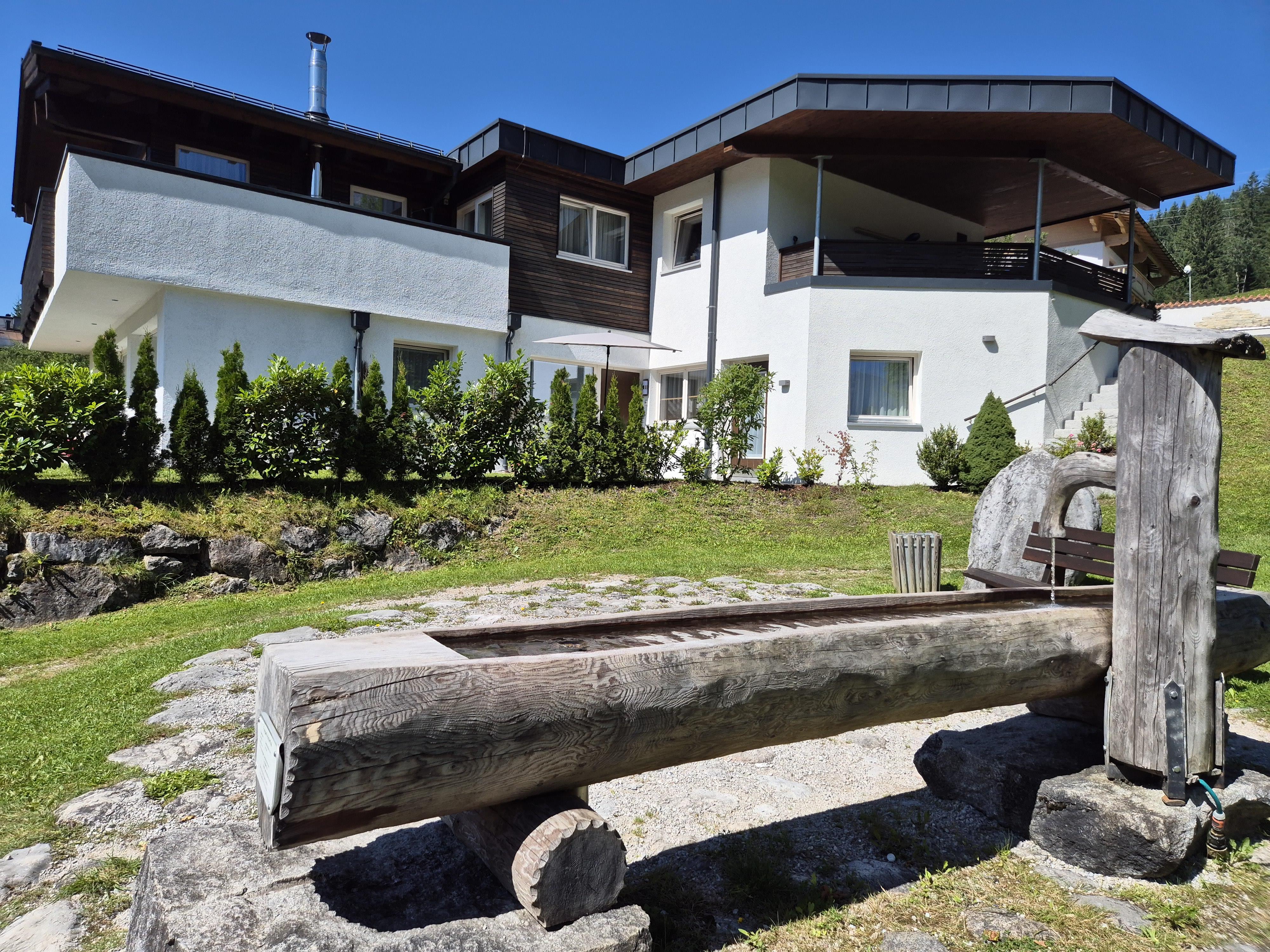 A modern house with a large balcony and a well-maintained green area. In the foreground, there is a rustic wooden bench.