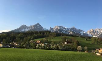 A picturesque landscape with green meadows and impressive mountains in the background. The sky is clear and radiant blue.