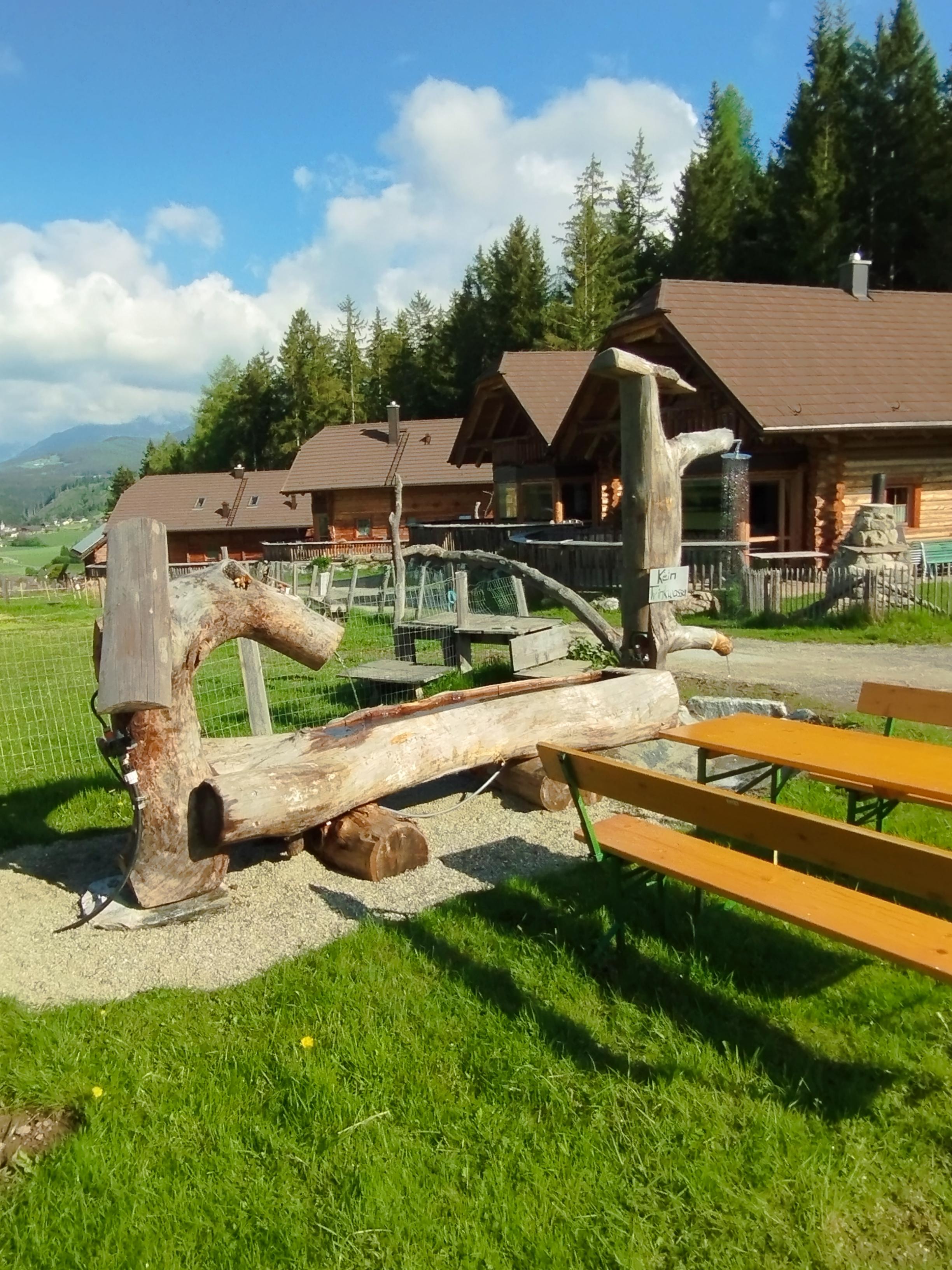 A rustic outdoor area with wooden seats and tables. In the background, there are cozy wooden cabins and a beautiful landscape with trees.