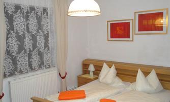 A cozy bedroom with a double bed and two pillows. There are light curtains and orange towels on the bed.