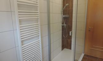 A modern bathroom with a glass shower cabin and a white towel radiator. The walls are light tiled and a wooden door leads outside.