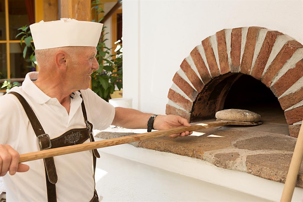 An elderly man in traditional clothing stands in front of a stone oven. He holds a wooden board to slide bread into the oven.