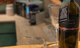 A bottle of wine is on a wooden table next to two glasses. In the background, a kitchen can be seen.