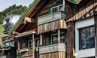 A traditional log cabin with wooden balconies and green window frames. In the background, gentle hills and a clear sky can be seen.