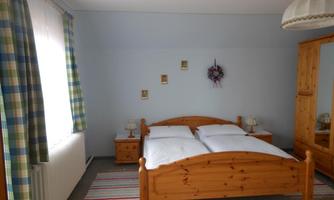 A cozy bedroom with a wooden double bed. The walls are light blue, and there are curtains with a checked pattern as well as a wooden camera.