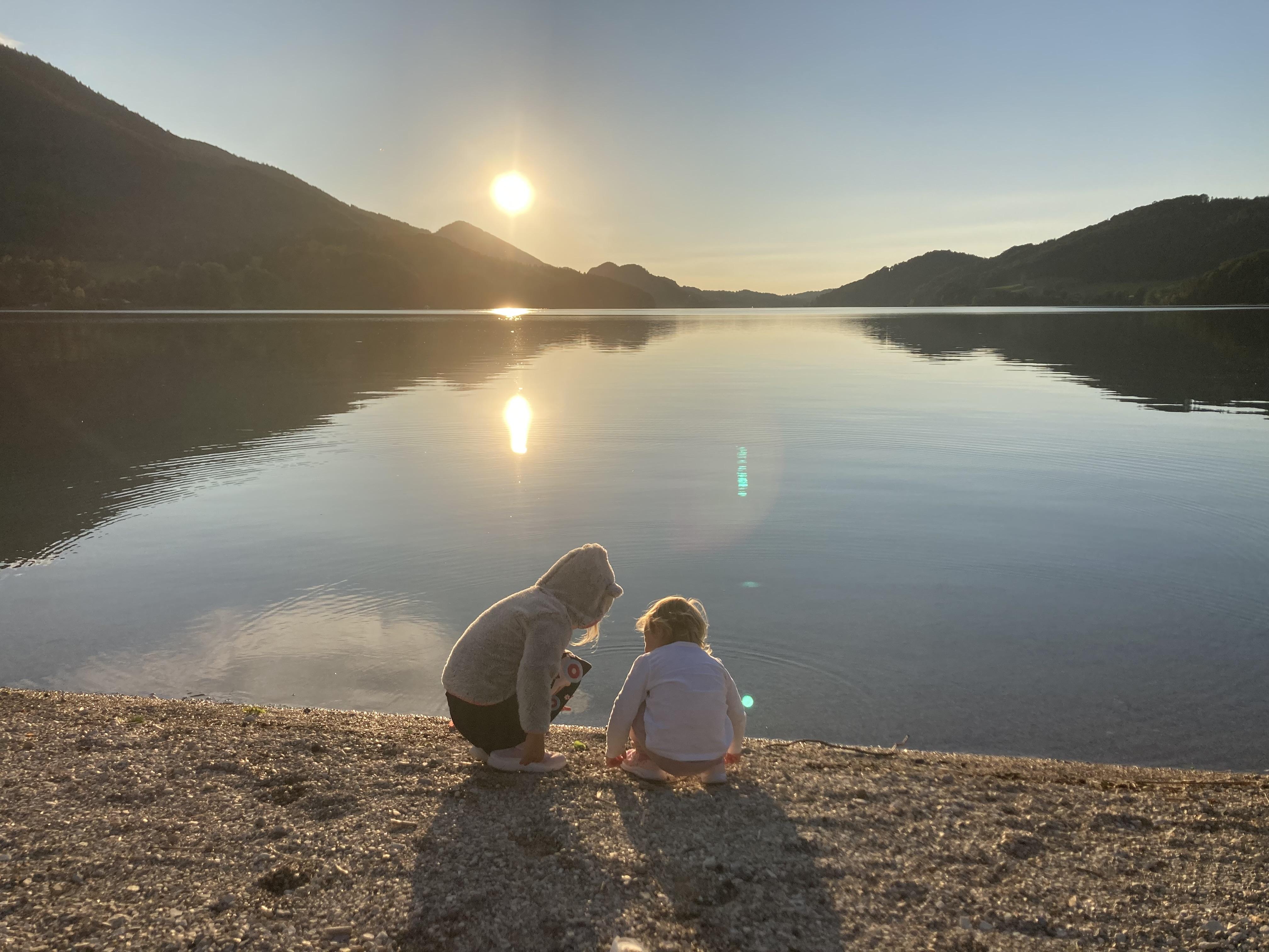 Two children are sitting by the shore of a tranquil lake, watching the sunset. The mountains are reflected in the water, and the atmosphere is peaceful.