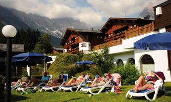 A group of people relaxes on reclining chairs in a garden. In the background, mountains and wooden houses can be seen under blue sun umbrellas.