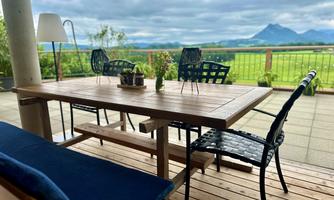 An inviting outdoor dining area with a wooden table and black chairs. In the background, green meadows and a mountain panorama can be seen.