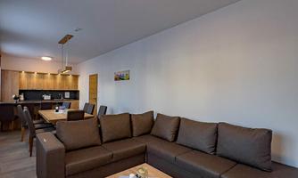 A modern living room with a brown sofa and a wooden table. In the background, a kitchenette is visible.