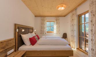 A cozy bedroom with wooden details, two beds, and colorful pillows. Large windows provide plenty of daylight and a view outside.