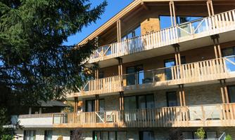 A modern building with wooden balconies and large windows. In the foreground stands a green tree and the sky is blue.
