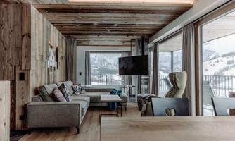 A modern living room with wood paneling and large windows. The view shows snow-covered mountains in the background.
