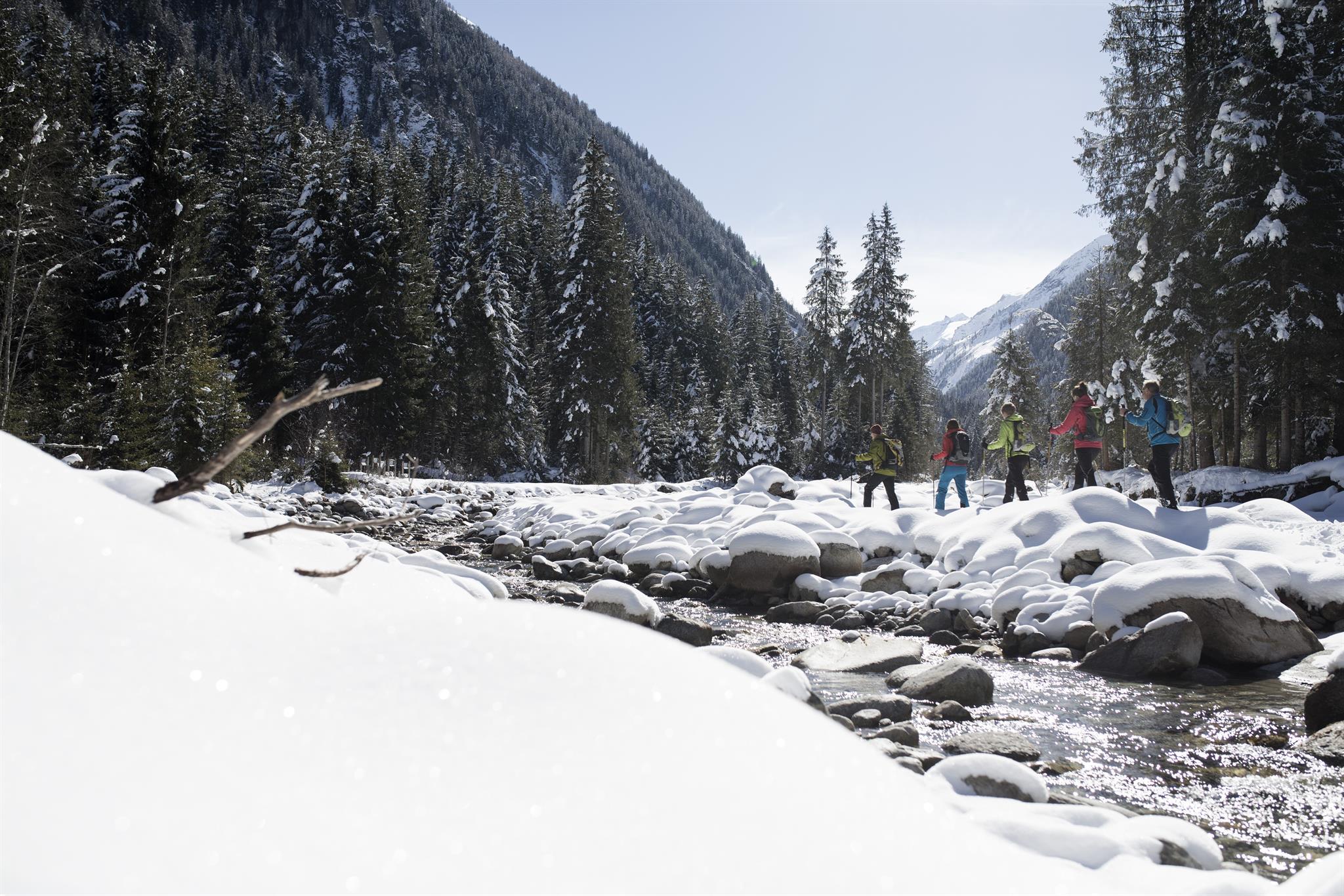 A group of people is hiking through a snowy landscape. In the foreground, a sparkling stream flows between snow-covered rocks.
