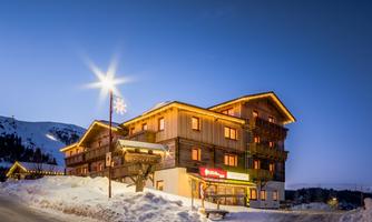 A cozy hotel in the mountains, surrounded by snow. The facade is illuminated and it is twilight.